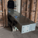Tracked carrier transporting multiple structural steel beams through a doorway during a tight‑access lift in Salamander Bay.